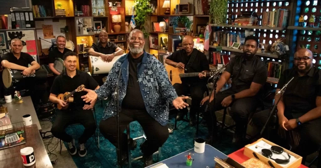 Uma banda de música brasileira se apresenta em um escritório ou estúdio repleto de estantes de livros, plantas e instrumentos musicais. O vocalista, Péricles, um homem negro de meia-idade com barba grisalha, está sentado no centro usando um casaco jeans, sorrindo e com os braços abertos. Ele está cercado por oito outros músicos, todos homens, a maioria vestindo camisas pretas, tocando bateria, violão, percussão e outros instrumentos.