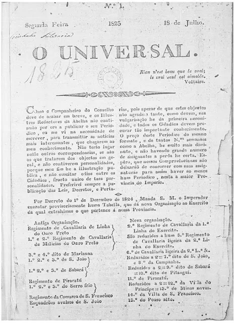 Uma fotografia em preto e branco de um exemplar da primeira edição do jornal histórico "O Universal", datado de 18 de Julho de 1825, Segunda-feira. O documento antigo apresenta-se com a tipografia da época, com o título "O UNIVERSAL" em destaque no topo, sob a epígrafe "N.º 1". O texto é organizado em colunas, com um pequeno trecho de Voltaire citado. A primeira coluna discorre sobre os objetivos do periódico. Abaixo, há uma seção intitulada "Antiga Organização" listando regimentos de cavalaria, e o documento possui marcas e anotações manuscritas claras no canto superior esquerdo.