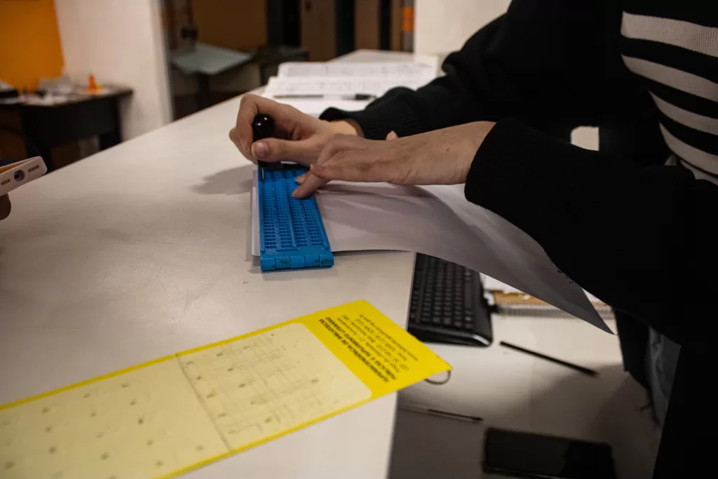 Um close-up de uma pessoa com vestimentas escuras e listradas no braço, utilizando uma reglete e um punção para escrever em Braille sobre uma folha de papel branco, que está apoiada em uma mesa de trabalho clara. O equipamento de escrita em Braille é uma reglete azul de plástico, e as mãos da pessoa estão centralizadas na imagem, em plena atividade. Em primeiro plano, no canto inferior, há um material amarelo com textos impressos, e ao fundo da mesa, partes de um teclado de computador e outros materiais de escritório podem ser vistos em um ambiente de iluminação mais baixa. A imagem ilustra o processo manual de produção de material acessível em Braille.
