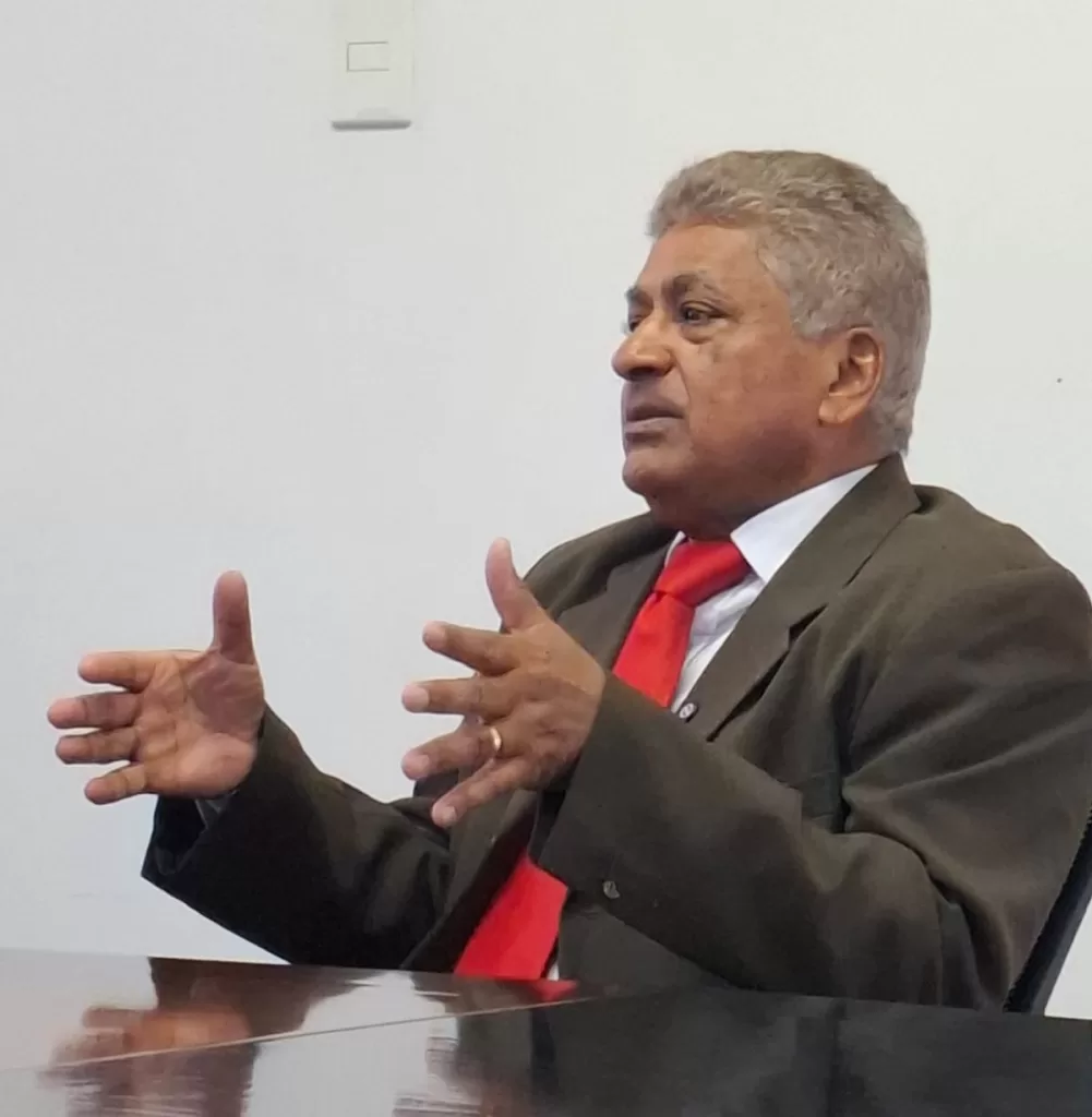 A imagem mostra Matias sentado à mesa, gesticulando com as mãos enquanto fala. Ele usa um terno escuro, camisa branca e gravata vermelha. Ele tem o cabelo grisalho curto e uma expressão séria, parecendo concentrado na conversa. O fundo é simples, com uma parede branca e um interruptor visível. Créditos: Sindifisco-MG/ Arquivo