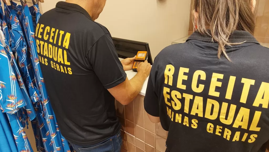 A imagem mostra dois agentes da Receita Estadual de Minas Gerais, identificados pelas camisetas pretas com letras amarelas. Eles estão em um ambiente interno do shopping, manuseando um equipamento eletrônico sobre um balcão. À esquerda, há roupas coloridas penduradas em cabides. A cena sugere uma ação de fiscalização ou verificação de documentos fiscais. Créditos: SEF MG/ Divulgação.