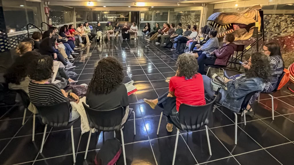 Uma fotografia noturna em um ambiente interno de uma biblioteca, mostrando uma Roda de Leitura ou debate. Um grupo diversificado de cerca de vinte pessoas está sentado em cadeiras pretas, dispostas em círculo em um piso de ladrilhos pretos brilhantes, participando de uma atividade. A iluminação ambiente é focada no centro do círculo, enquanto o fundo permanece mais escuro. À direita, um grande objeto artístico, que se assemelha à cabeça de um dinossauro ou ser mitológico marrom-escuro, está parcialmente visível. As janelas no fundo refletem a luz e mostram o exterior escuro, indicando ser um evento noturno.