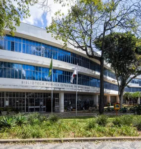 Em uma foto externa, tirada em um dia ensolarado, vê-se a fachada de um grande edifício moderno, branco e curvo, de vários andares: a Biblioteca Pública Estadual de Minas Gerais, como indicado pelo texto em letras maiúsculas na fachada. O prédio possui longas faixas horizontais de janelas de vidro azulado que refletem o céu azul e algumas nuvens brancas. Em primeiro plano, há um jardim com plantas verdes variadas, como agaves, e grama um pouco seca. Duas bandeiras, uma do Brasil e outra que parece ser de Minas Gerais (vermelha e branca), estão hasteadas em mastros em frente à entrada. Árvores grandes e frondosas, com folhas verdes, estão presentes no pátio, uma delas em destaque à direita, projetando sombras no prédio e no chão sob a luz solar intensa.