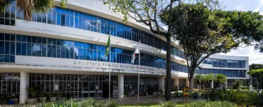 Em uma foto externa, tirada em um dia ensolarado, vê-se a fachada de um grande edifício moderno, branco e curvo, de vários andares: a Biblioteca Pública Estadual de Minas Gerais, como indicado pelo texto em letras maiúsculas na fachada. O prédio possui longas faixas horizontais de janelas de vidro azulado que refletem o céu azul e algumas nuvens brancas. Em primeiro plano, há um jardim com plantas verdes variadas, como agaves, e grama um pouco seca. Duas bandeiras, uma do Brasil e outra que parece ser de Minas Gerais (vermelha e branca), estão hasteadas em mastros em frente à entrada. Árvores grandes e frondosas, com folhas verdes, estão presentes no pátio, uma delas em destaque à direita, projetando sombras no prédio e no chão sob a luz solar intensa.