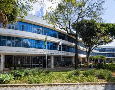 Em uma foto externa, tirada em um dia ensolarado, vê-se a fachada de um grande edifício moderno, branco e curvo, de vários andares: a Biblioteca Pública Estadual de Minas Gerais, como indicado pelo texto em letras maiúsculas na fachada. O prédio possui longas faixas horizontais de janelas de vidro azulado que refletem o céu azul e algumas nuvens brancas. Em primeiro plano, há um jardim com plantas verdes variadas, como agaves, e grama um pouco seca. Duas bandeiras, uma do Brasil e outra que parece ser de Minas Gerais (vermelha e branca), estão hasteadas em mastros em frente à entrada. Árvores grandes e frondosas, com folhas verdes, estão presentes no pátio, uma delas em destaque à direita, projetando sombras no prédio e no chão sob a luz solar intensa.