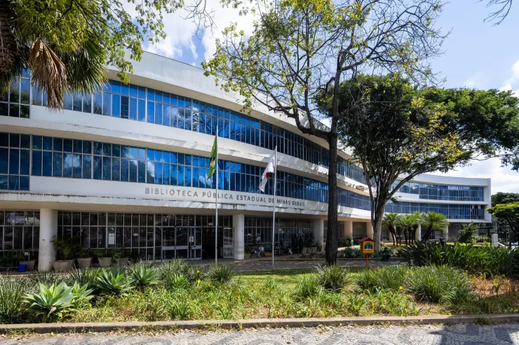 Em uma foto externa, tirada em um dia ensolarado, vê-se a fachada de um grande edifício moderno, branco e curvo, de vários andares: a Biblioteca Pública Estadual de Minas Gerais, como indicado pelo texto em letras maiúsculas na fachada. O prédio possui longas faixas horizontais de janelas de vidro azulado que refletem o céu azul e algumas nuvens brancas. Em primeiro plano, há um jardim com plantas verdes variadas, como agaves, e grama um pouco seca. Duas bandeiras, uma do Brasil e outra que parece ser de Minas Gerais (vermelha e branca), estão hasteadas em mastros em frente à entrada. Árvores grandes e frondosas, com folhas verdes, estão presentes no pátio, uma delas em destaque à direita, projetando sombras no prédio e no chão sob a luz solar intensa.