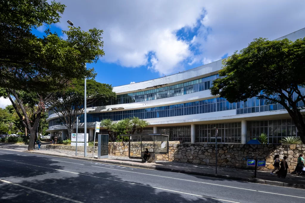 
Uma vista lateral da Biblioteca Pública Estadual de Minas Gerais, com sua fachada curva e horizontal de cor branca, que se estende sob um céu parcialmente nublado e azul. A parte superior do edifício exibe longas faixas de janelas azuis refletivas, enquanto o andar térreo possui pilares finos. A base da construção é cercada por um muro rústico feito de pedras claras e irregulares. Em primeiro plano, uma rua asfaltada escura e, à direita, uma calçada arborizada onde se vê um ponto de ônibus com uma pessoa sentada na área de espera, e duas pessoas caminhando mais ao fundo. Postes de iluminação e copas de árvores grandes e verdes emolduram a cena nos cantos esquerdo e direito.