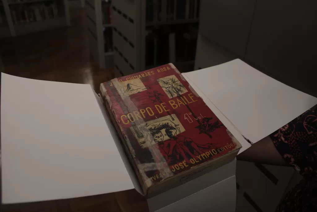 Um plano detalhado foca no livro "Corpo de Baile", de João Guimarães Rosa, uma edição antiga em capa dura vermelha e amarelada, com o texto e ilustrações em preto e branco. O livro está ligeiramente aberto e posicionado sobre uma superfície escura, com duas folhas brancas abertas ao lado, em contraste com a capa. O ambiente, que parece ser um setor de acervo da biblioteca, é escuro, com iluminação focal no objeto principal. No fundo desfocado, à esquerda e no centro, é possível distinguir estantes escuras e altas repletas de livros, sugerindo um vasto volume de obras históricas.