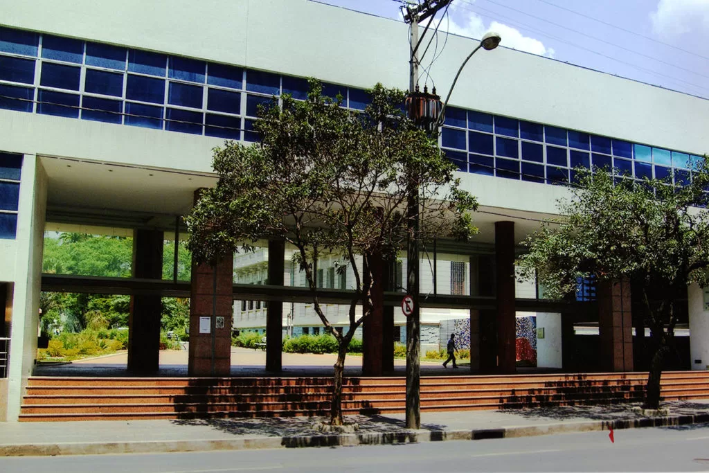 Uma vista da entrada principal de um edifício público de arquitetura moderna e horizontal, com fachada superior branca e longas janelas refletivas azuis. A entrada é marcada por um grande pórtico sustentado por colunas largas e escuras, com vigas horizontais que criam uma área de sombra. Em frente ao pórtico, há uma escadaria ampla na cor laranja-tijolo, que se estende por toda a largura da fachada. No primeiro plano, uma rua asfaltada e uma calçada, com uma árvore de tronco fino e galhos modestos centralizada. Um poste de iluminação com fiação proeminente está à direita da árvore, e uma pessoa de longe caminha sob o pórtico.