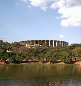 Imagem panorâmica da Lagoa da Pampulha em Belo Horizonte, com o Ginásio Mineirinho ao fundo, vegetação nas margens e céu azul, representando a revitalização da região e o projeto de retomada da navegação.