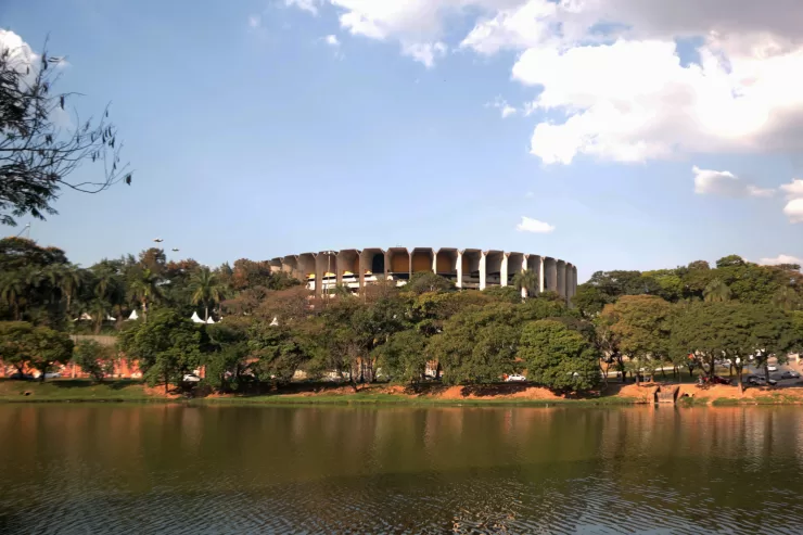 Imagem panorâmica da Lagoa da Pampulha em Belo Horizonte, com o Ginásio Mineirinho ao fundo, vegetação nas margens e céu azul, representando a revitalização da região e o projeto de retomada da navegação.