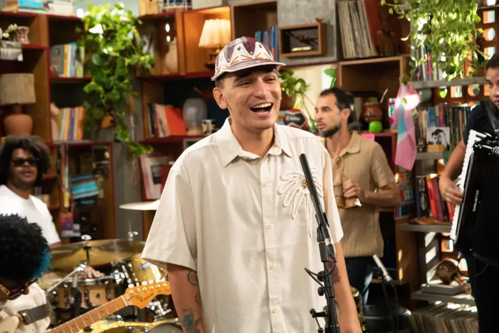 Um homem, com um chapéu estampado e camisa bege bordada, canta sorridente em um microfone.  Ao fundo, outros músicos são parcialmente visíveis, incluindo um homem de óculos escuros e cabelo crespo, um baterista e um músico tocando acordeão. O cenário é um estúdio com prateleiras cheias de livros e plantas.