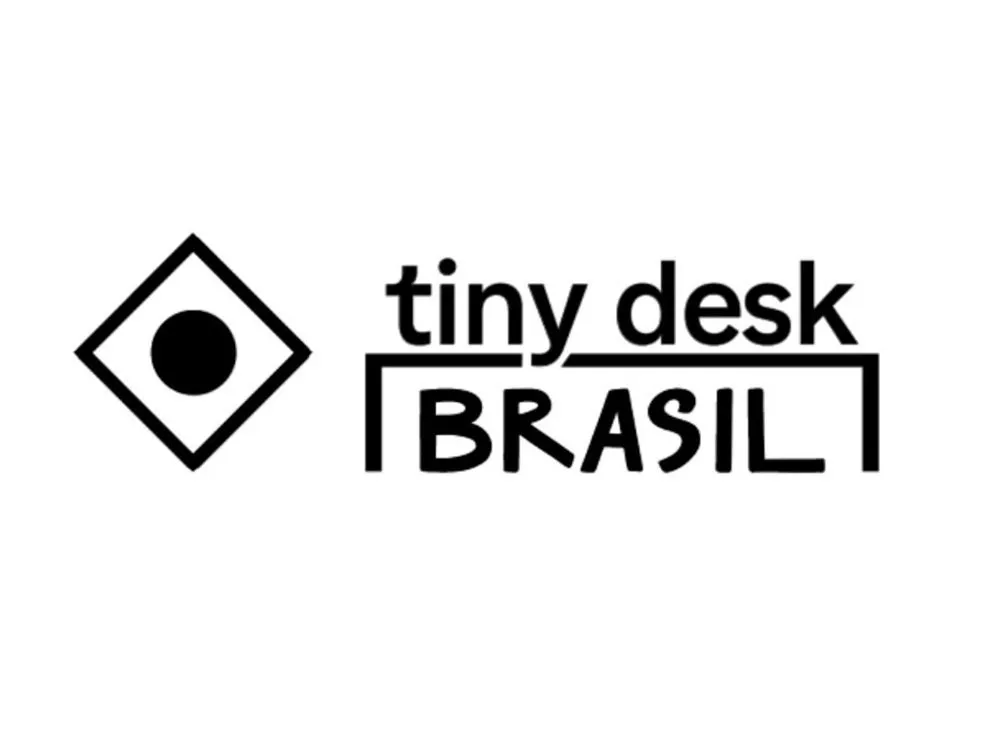 O logotipo do "Tiny Desk Brasil". À esquerda, um quadrado preto com um círculo branco no centro fazendo referência a bandeira do Brasil. À direita, as palavras "tiny desk" em fonte minúscula, com "BRASIL" em letras maiúsculas e sublinhadas abaixo, tudo em preto sobre um fundo branco.