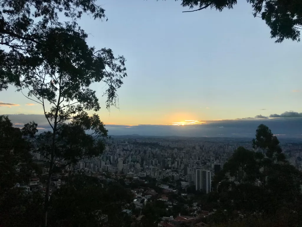 A imagem mostra o horizonte de prédios em Belo Horizonte. Ao fundo, o sol está se pondo. Galhos de árvores ocupam as laterais da imagem.