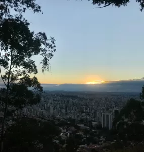 A imagem mostra o horizonte de prédios em Belo Horizonte. Ao fundo, o sol está se pondo. Galhos de árvores ocupam as laterais da imagem.