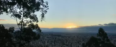 A imagem mostra o horizonte de prédios em Belo Horizonte. Ao fundo, o sol está se pondo. Galhos de árvores ocupam as laterais da imagem.