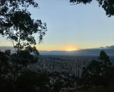 A imagem mostra o horizonte de prédios em Belo Horizonte. Ao fundo, o sol está se pondo. Galhos de árvores ocupam as laterais da imagem.