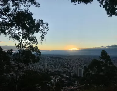 A imagem mostra o horizonte de prédios em Belo Horizonte. Ao fundo, o sol está se pondo. Galhos de árvores ocupam as laterais da imagem.