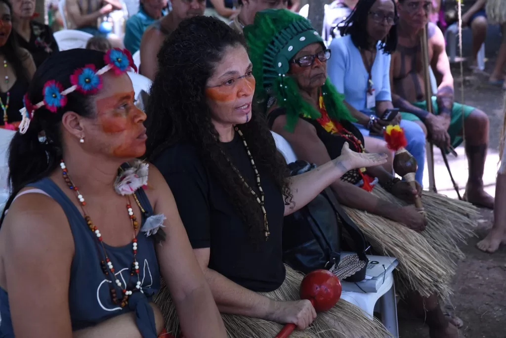 A imagem mostra um grupo indígena reunido ao ar livre. Em destaque, três mulheres com pinturas faciais e adornos tradicionais participam ativamente, segurando maracás. Uma delas fala enquanto gesticula, e ao fundo outras pessoas observam.
