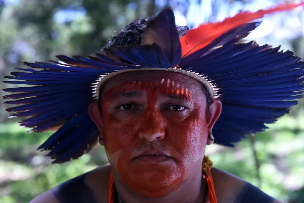 Um homem indígena, com a feição seria usando um cocar azul e com o rosto pintado de laranja. Com fundo desfocado