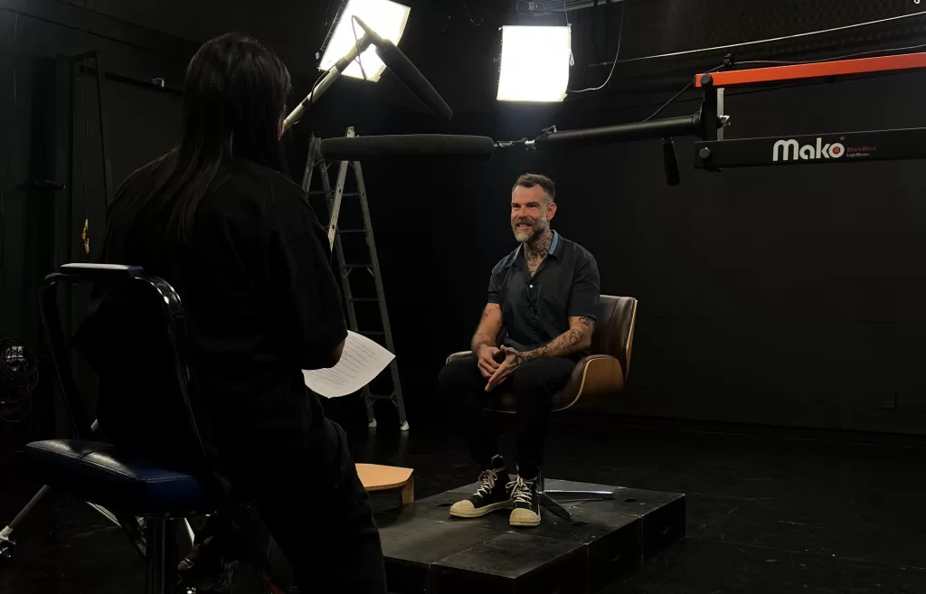 Em um estúdio de gravação, o entrevistado fala sobre o processo de apuração e os bastidores de suas reportagens. A cena é iluminada por refletores e acompanhada de equipamentos profissionais de som, revelando o clima de concentração e cuidado característico de produções jornalísticas que buscam contar histórias reais com profundidade.