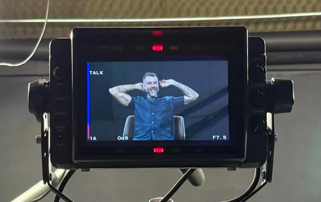 A fotografia captura o clima leve dos bastidores de uma produção audiovisual. No monitor da câmera, o entrevistado surge relaxado,se ajustando e sorrindo. O registro mostra um raro instante de pausa durante uma gravação marcada por conversas intensas sobre jornalismo e narrativa.