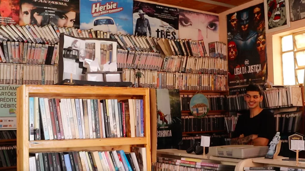 Interior do Ponto de Cultura Capim Branco, repleto de livros, DVDs e pôsteres, com um atendente atrás do balcão.