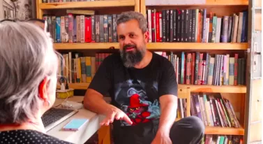 Repórter Katia Torres conversa com o agente cultural Andre Serra dentro do Ponto De cultura Capim Branco, com estantes cheias de livros e filmes ao fundo.