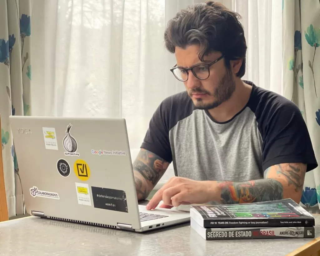 Na imagem, um homem branco sentado na frente de um notebook cinza que possui vários adesivos na parte de cima. Ao lado, dois livros compondo o cenário. No fundo, uma cortina branca deixa a luz do sol entrar, refletindo no ambiente