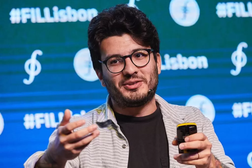 Um homem branco está ao centro da imagem, dando uma palestra. Ele usa óculos de armação preta e veste uma camisa preta e uma blusa social listrada aberta por cima. Está olhando para o lado e gesticulando com as mãos.