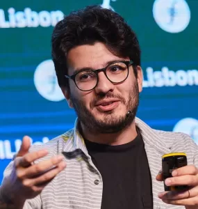Um homem branco está ao centro da imagem, dando uma palestra. Ele usa óculos de armação preta e veste uma camisa preta e uma blusa social listrada aberta por cima. Está olhando para o lado e gesticulando com as mãos.