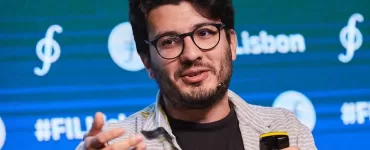 Um homem branco está ao centro da imagem, dando uma palestra. Ele usa óculos de armação preta e veste uma camisa preta e uma blusa social listrada aberta por cima. Está olhando para o lado e gesticulando com as mãos.