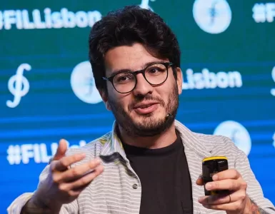 Um homem branco está ao centro da imagem, dando uma palestra. Ele usa óculos de armação preta e veste uma camisa preta e uma blusa social listrada aberta por cima. Está olhando para o lado e gesticulando com as mãos.