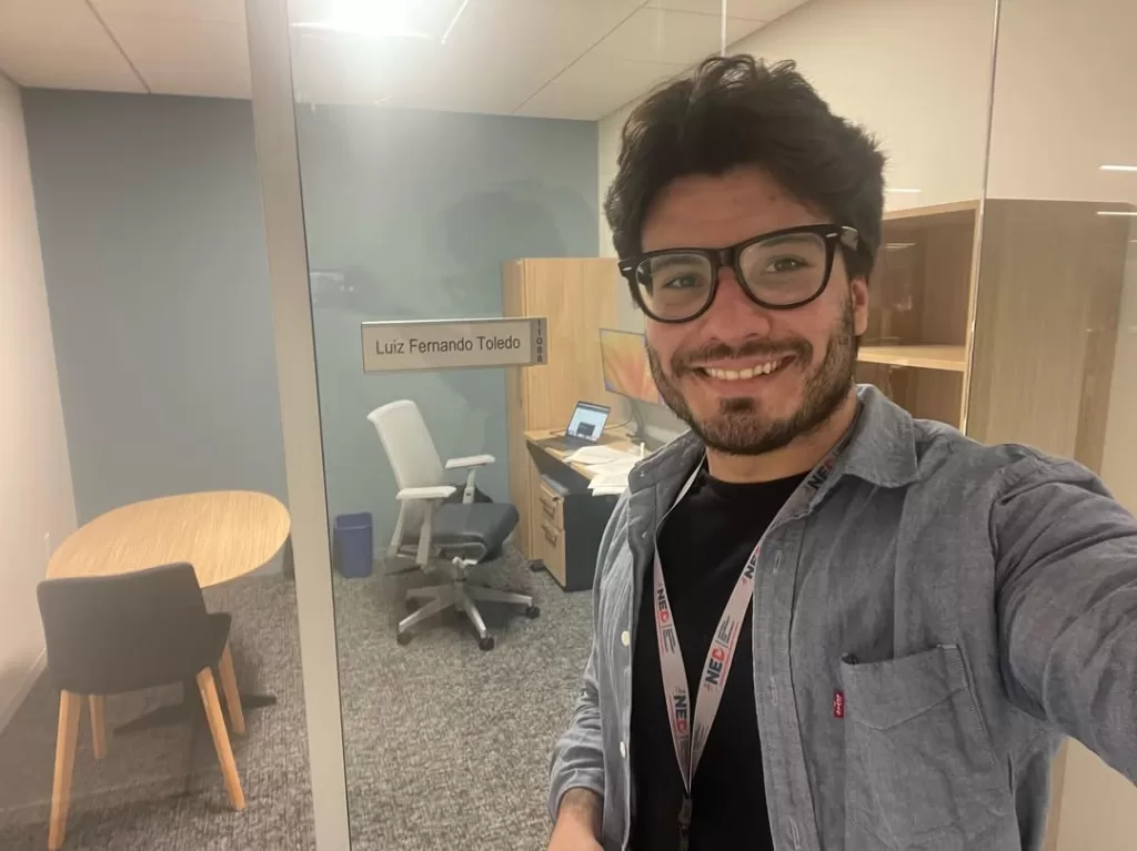Um homem branco está tirando uma selfie ao lado de uma placa com seu nome "Luiz Fernando Toledo" na porta da sala. Com parede de vidro, ao fundo tem uma mesa com dois computadores e uma cadeira de trabalho e outra mesa de madeira na mesma sala.