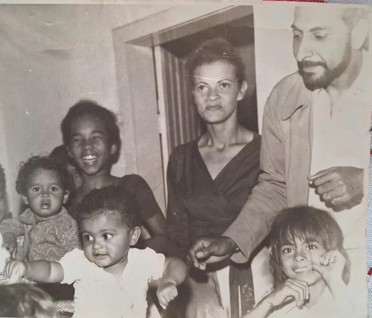 Na fotografia em preto e branco, Zu Moreira aparece em primeiro plano, com o braço estendido, cercada por familiares e amigos. À direita, seu irmão no colo, enquanto os pais observam a cena ao fundo e os convidados celebram o momento.