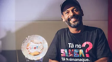 Zu Moreira em primeiro plano usando boné e vestindo uma camisa preta com a estampa do Almanaque do Samba, ao seu lado um prato decorativo do Almanaque do Samba e ao fundo uma parede branca.