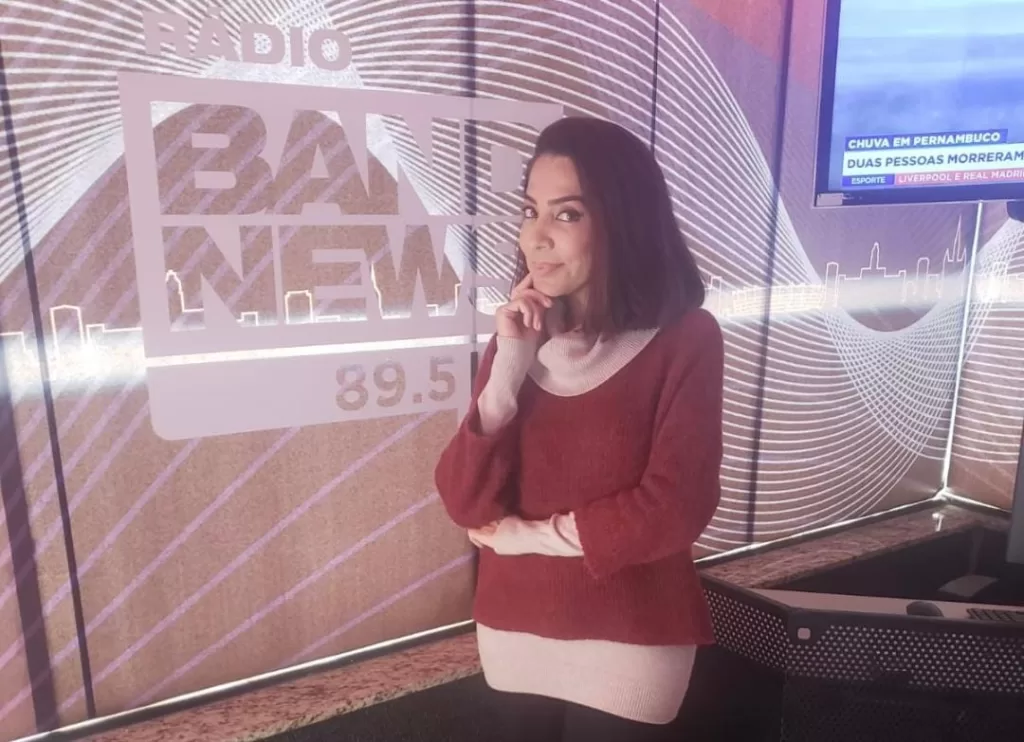 Foto colorida, em primeiro plano, Vianna está com uma mão na altura
da cintura e outra tocando o queixo, posando em frente o cenário de gravações da
Band News. Em segundo plano é possível ver a placa da rádio onde está escrito
“Rádio Band News 89.5” iluminada por uma luz branca.