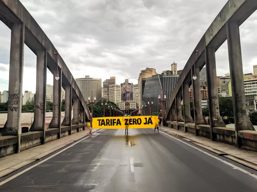 Uma faixa escrito "Tarifa Zero já" ocupa o centro da imagem. Duas pessoas seguram a faixa no meio da rua do viaduto Santa Tereza, em Belo Horizonte