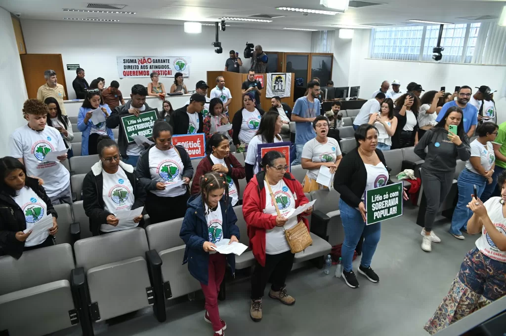 Uma fotografia em ângulo aberto e nível dos olhos capta um grupo de pessoas em pé, lotando as primeiras filas de cadeiras de um auditório que parece ser uma sala de reuniões ou câmara municipal.
A maioria das pessoas está virada para a frente da sala, embora algumas estejam viradas para os lados ou para trás. Muitos estão usando camisetas brancas, algumas delas com uma estampa verde na frente (parecendo um símbolo ou mapa), e vários indivíduos, especialmente nas filas da frente, estão usando babadores ou coletes brancos sobre a camiseta com uma estampa semelhante ou idêntica.