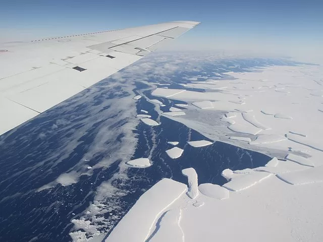Vista aérea tirada da janela de um avião, mostrando parte da asa branca da aeronave no canto superior esquerdo. Abaixo, observa-se uma paisagem polar com o mar coberto por grandes placas de gelo brancas e fragmentadas flutuando sobre a água azul-escura.