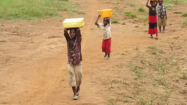 Foto de quatro crianças negras caminhando por uma estrada de terra batida com vegetação rasteira. Em primeiro plano, duas crianças carregam grandes galões amarelos equilibrados sobre a cabeça. Ao fundo, outras duas crianças observam paradas