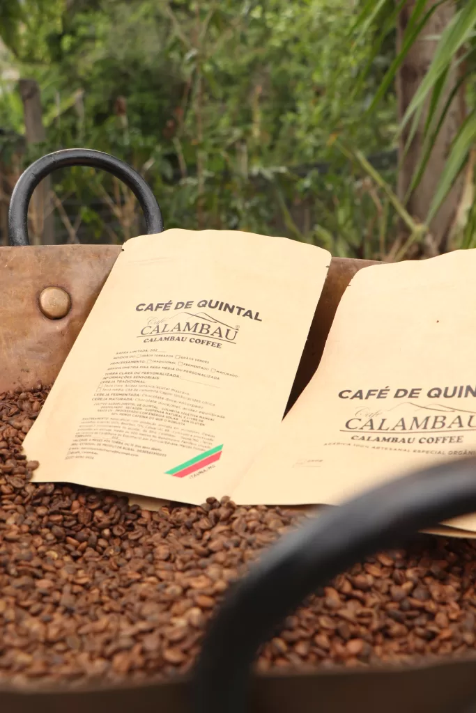 Dois pacotes de café artesanal apoiados sobre muitos grãos de café torrados. Nos rótulos lê-se “Café de Quintal – Calambau Coffee”. Ao fundo, vegetação verde desfocada, sugerindo um ambiente rural.