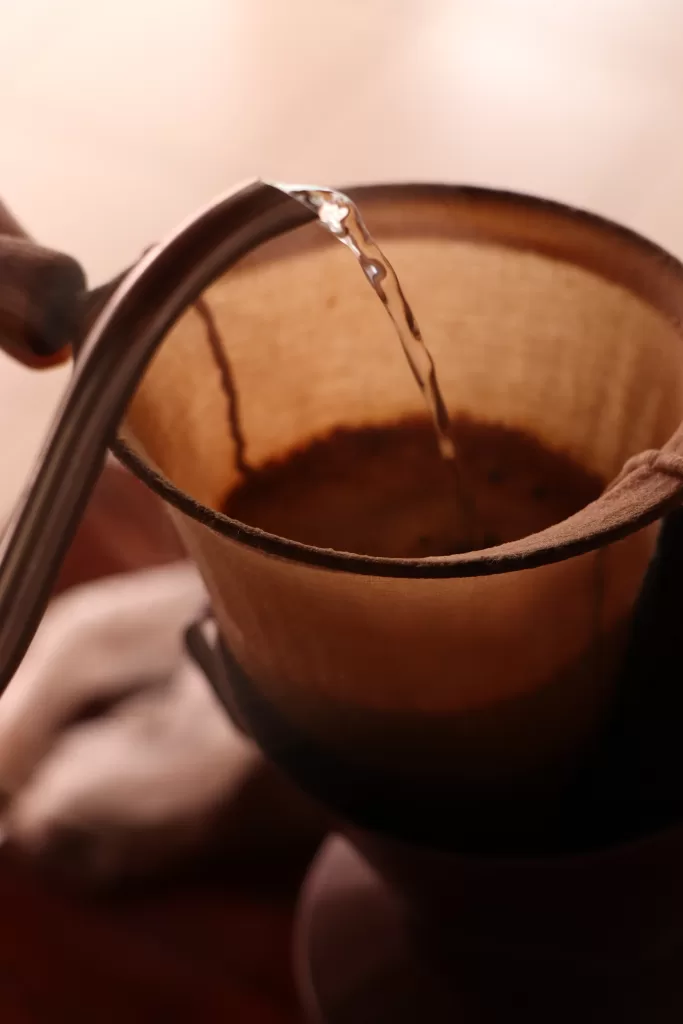 Close em um filtro de pano com café moído. Água quente é despejada lentamente por um bico de chaleira, iniciando o preparo do café. A cena tem tons quentes e atmosfera aconchegante.