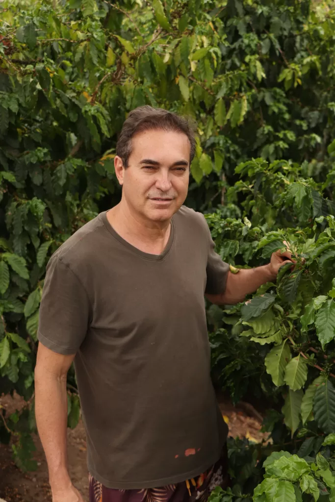 Homem adulto, de cabelos curtos e escuros, em pé entre pés de café carregados de folhas verdes. Ele usa camiseta de manga curta e segura um galho da planta, olhando para a câmera. O ambiente é uma lavoura de café ao ar livre.