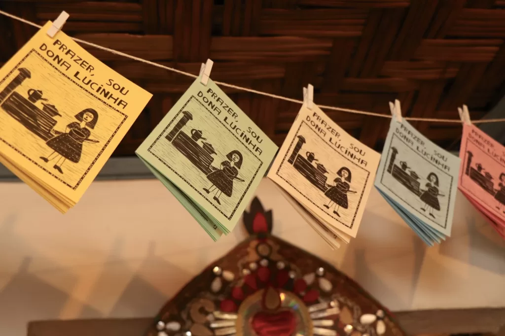 Cartões pequenos e coloridos pendurados em um barbante por prendedores de roupa. Os cartões são amarelos, verdes, azuis e rosados, cada um com uma ilustração simples em preto de uma mulher ao lado de um fogão a lenha. No topo de cada cartão está escrito “Prazer, sou Dona Lucinha”. Ao fundo, aparece uma parede de palha trançada e, mais abaixo, um objeto decorativo em formato de coração com detalhes vermelhos e dourados.