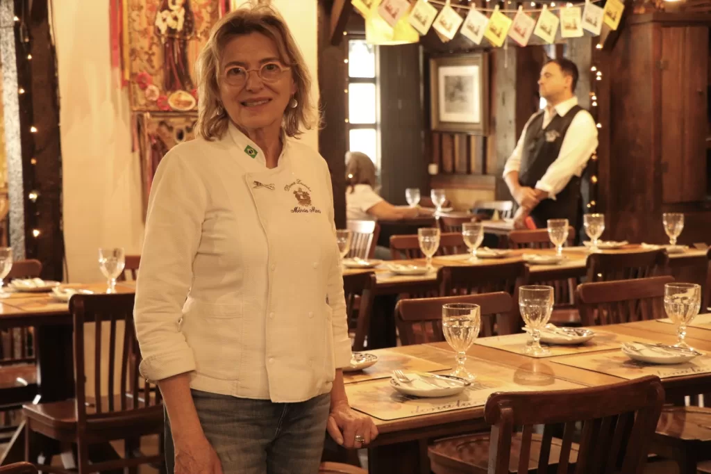 Mulher adulta, de cabelos claros e curtos, usa óculos e veste um dólmã branco de chef. Ela está em um restaurante rústico, com mesas de madeira postas com copos e pratos. Ao fundo, outras pessoas e uma decoração acolhedora com luzes quentes.