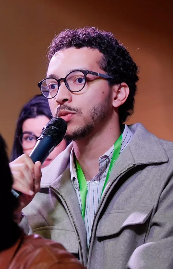 Foto de um jovem adulto, homem, de pele clara e cabelo escuro e cacheado, com barba rala e bigode. Ele usa óculos de aro preto e está falando em um microfone, que segura com a mão esquerda. Ele veste uma camisa social, sobreposta por uma jaqueta cinza-claro, com zíper frontal. Um crachá verde-limão com a inscrição "Google" está pendurado em seu pescoço. O homem tem uma expressão concentrada, com a boca ligeiramente aberta enquanto fala. No fundo, à esquerda e ligeiramente desfocada, aparece parcialmente uma segunda pessoa, uma mulher de cabelo escuro, com óculos, olhando para frente.