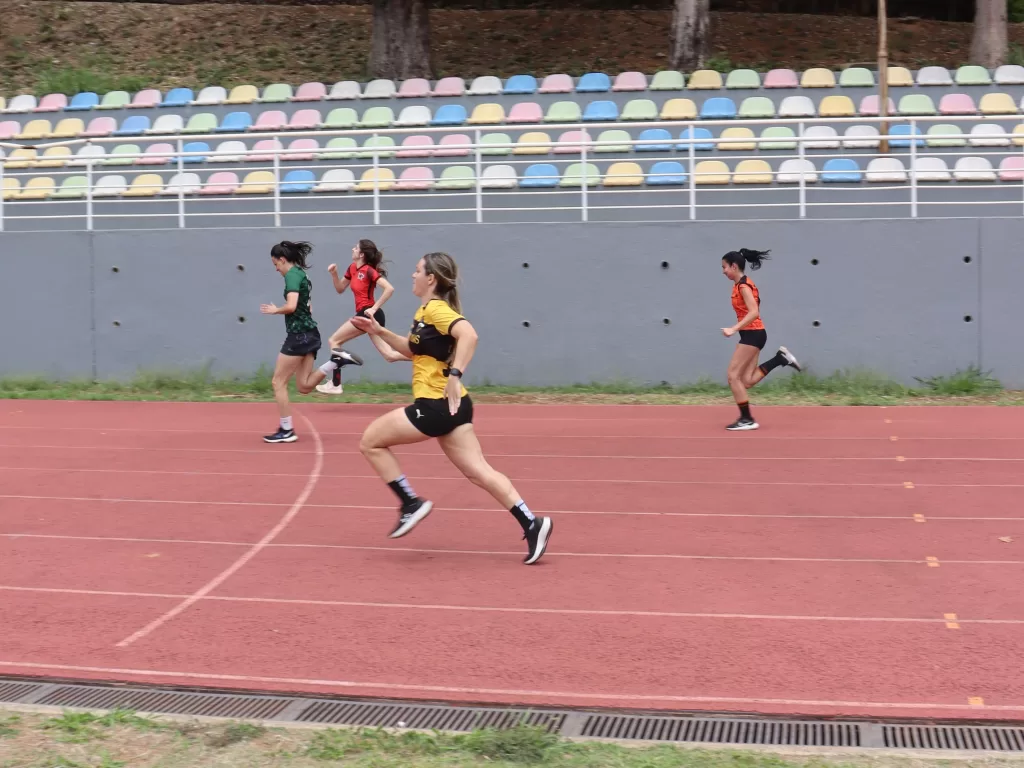 Foto horizontal de uma corrida em pista de atletismo ao ar livre. Quatro mulheres correm em raias paralelas. No centro, uma atleta com camiseta amarela e shorts pretos aparece à frente, em um grande passo, com um dos pés no ar. À esquerda, duas corredoras vestem verde e vermelho; à direita, outra atleta usa camiseta laranja. A pista é vermelha, com marcações brancas, e ao fundo há arquibancadas vazias com assentos coloridos.