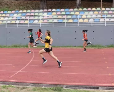 Foto horizontal de uma corrida em pista de atletismo ao ar livre. Quatro mulheres correm em raias paralelas. No centro, uma atleta com camiseta amarela e shorts pretos aparece à frente, em um grande passo, com um dos pés no ar. À esquerda, duas corredoras vestem verde e vermelho; à direita, outra atleta usa camiseta laranja. A pista é vermelha, com marcações brancas, e ao fundo há arquibancadas vazias com assentos coloridos.