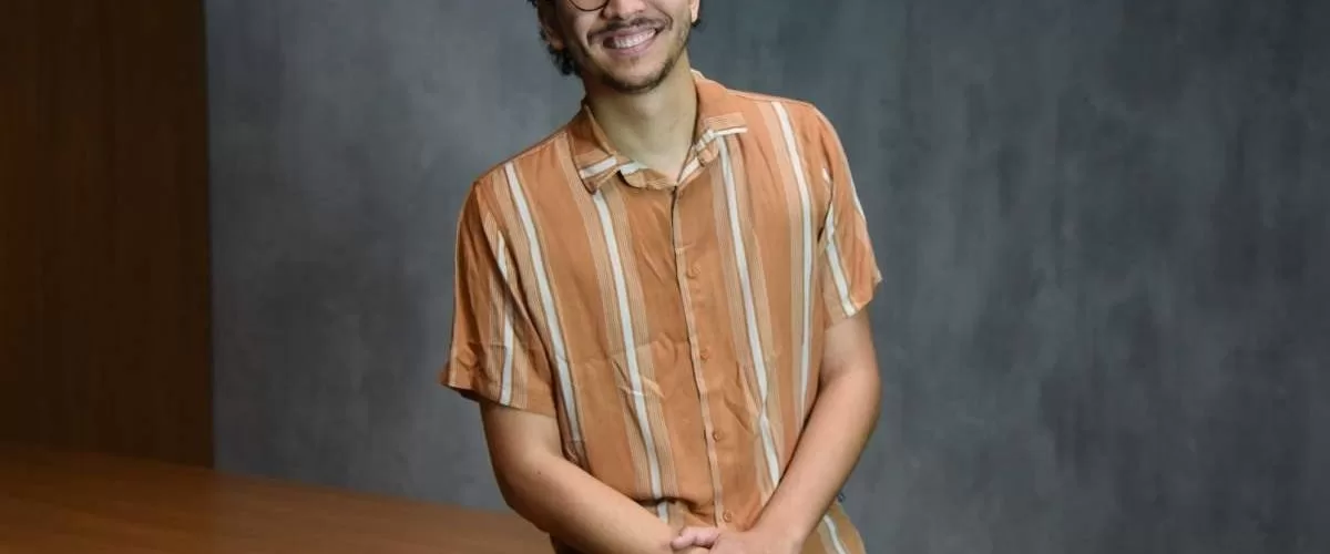 Foto de estúdio, à meia altura, de um jovem adulto, homem, de pele clara e cabelo escuro cacheado, com barba rala, usando óculos de aro fino e preto. Ele está sentado em uma mesa. O homem veste uma camisa casual de botões, de manga curta, na cor laranja com listras finas em um tom mais claro, e calça jeans azul-claro. Ele está sorrindo, olhando diretamente para a câmera, com as mãos cruzadas e apoiadas no colo. O fundo é uma parede cinza-escura, simples, com iluminação de estúdio.